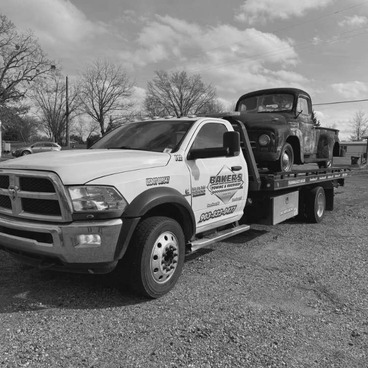 Flatbed tow truck transporting a vintage step-side pickup truck on gravel surface